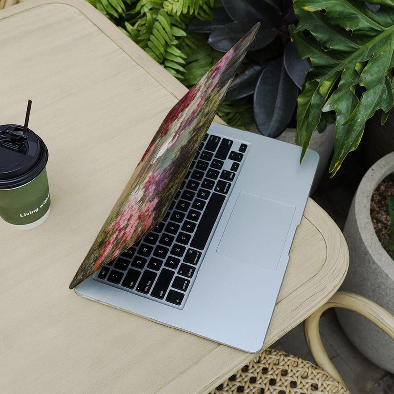 May include: A silver laptop with a black keyboard and a colorful floral design on the lid sits on a light wood table. A green coffee cup with the text "Living with" is next to the laptop. Plants are in the background.