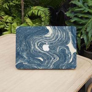 Op de afbeelding: Een laptop met een marineblauw en crèmekleurig wervelend patroon. Het Apple-logo is in het midden zichtbaar. De laptop staat op een lichtgekleurde houten tafel, met groene bladeren op de achtergrond.