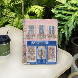 May include: A decorative book-shaped box featuring a detailed illustration of a quaint book shop. The shop has a pink facade with blue accents, flower boxes, and the text "BOOK SHOP" above the entrance. The box is on a light-colored surface.