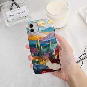 May include: A smartphone case featuring a colourful stained glass style desert landscape illustration. The design depicts tall green cacti, smaller succulents, and rolling hills in shades of blue, purple, orange, and yellow under a cloudy sky.