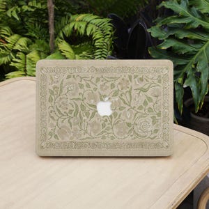 Könnte beinhalten: Eine beige Laptop-Hülle mit einem floralen Muster in Grün- und Beigetönen. Die Hülle hat ein detailliertes Design aus Blumen und Blättern, mit dem Apple-Logo in der Mitte. Die Hülle liegt auf einem hellen Holztisch.