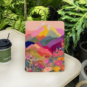 Puede incluir: Una funda rectangular para tableta con una ilustración vibrante y multicolor de cadenas montañosas en capas en tonos rosa, amarillo, verde y morado, con un primer plano lleno de flores silvestres detalladas y coloridas y elementos botánicos.