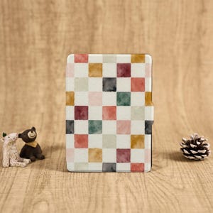 Pode incluir: Uma capa retangular para tablet com um padrão xadrez colorido. O padrão apresenta quadrados em tons de vermelho, rosa, amarelo, verde e cinza sobre um fundo branco. A capa está fechada e tem uma borda preta.
