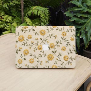 May include: A laptop hard shell case featuring a cream-colored fabric texture with an embroidered pattern of yellow flowers and green vines. The design includes a central cutout revealing the silver Apple logo on the laptop surface.