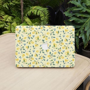 May include: A laptop with a yellow cover. The cover has a watercolour pattern of lemons, green leaves, and small blue berries. The laptop has a white Apple logo.