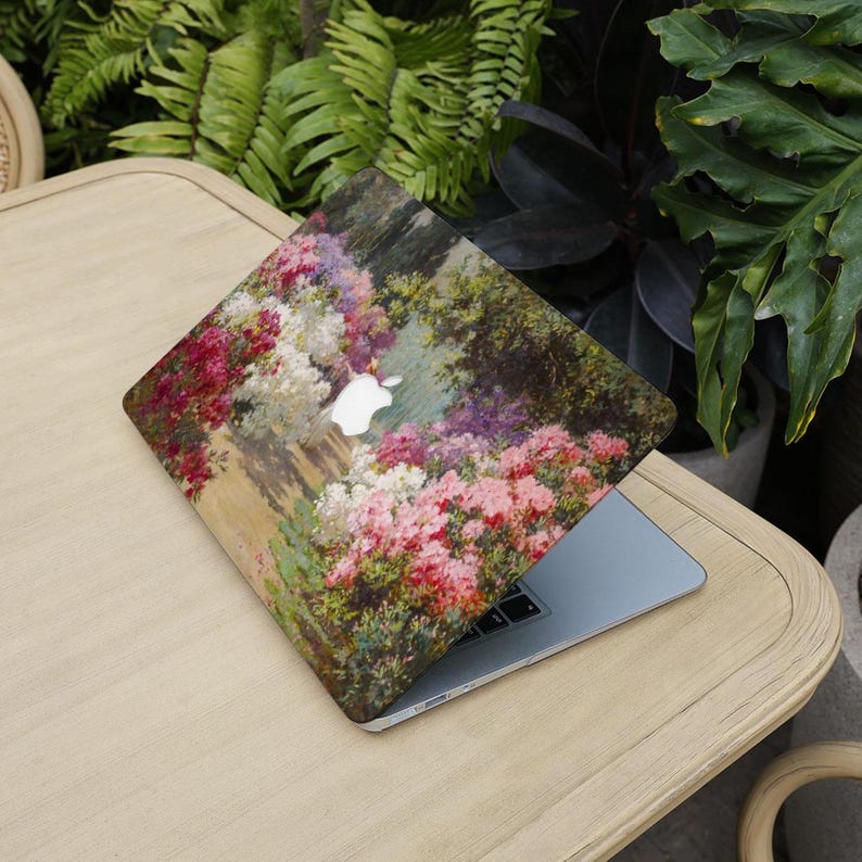 May include: A laptop with a floral design case featuring a painting of a garden path with pink, white, and purple flowers. The laptop is open on a light wood table, surrounded by green plants.