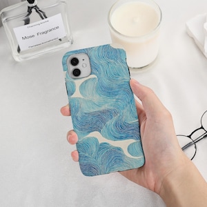 May include: A smartphone case featuring a blue and white wave design. The case has a shiny surface and is held in a hand. The phone case is designed for a specific phone model. The background includes a perfume bottle and a candle.