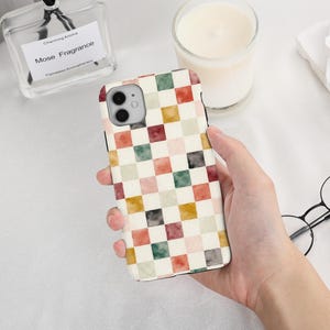 May include: A smartphone case featuring a repeating checkered pattern of watercolor-style squares in muted shades of green, red, yellow, and grey on a cream background, with a cutout for a dual-lens camera system on the upper left corner.