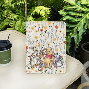 Może przedstawiać: Etui na tablet z kwiatowym wzorem z żółtych, pomarańczowych i niebieskich kwiatów. Etui przedstawia kreskówkową ilustrację Kubusia Puchatka i jego przyjaciół. Etui leży na jasnym drewnianym stole z filiżanką kawy.