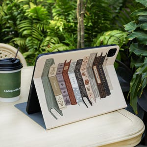 May include: An open tablet case featuring a design of a row of vintage books in various colors. The books have titles such as "Evermore" and "Midnights". A coffee cup with the text "Living with" is in the background.