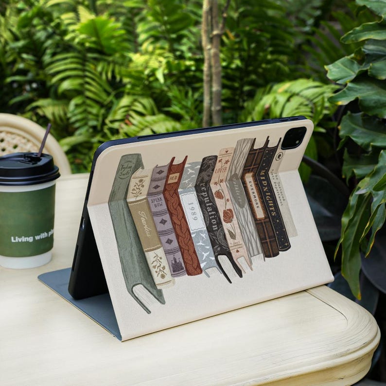 May include: An open tablet case featuring a design of a row of vintage books in various colors. The books have titles such as "Evermore" and "Midnights". A coffee cup with the text "Living with" is in the background.