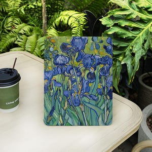 May include: A tablet case featuring a painting of blue irises in a field, with green and yellow accents. The artwork is reminiscent of Van Gogh's style. A coffee cup with the text "Living with" is on the left.