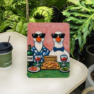 May include: A rectangular painting featuring two white geese wearing black sunglasses and sitting at a table. One goose wears a blue patterned shirt, the other a blue bow tie. A pizza box, wine glasses, a bottle labeled PINK, and food are on the table.