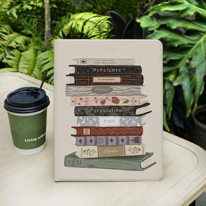 May include: A beige tablet case featuring a stack of books with titles like "Midnights," "Evermore," and "Reputation." The case has a book-like design with a coffee cup to the left. The cup is green with the words "Living with" on it.