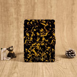 May include: A rectangular e-reader case featuring a black and amber tortoiseshell pattern with the name CECILIA printed in gold lettering at the bottom center. The case has a smooth finish and a small tab on the right side for opening.
