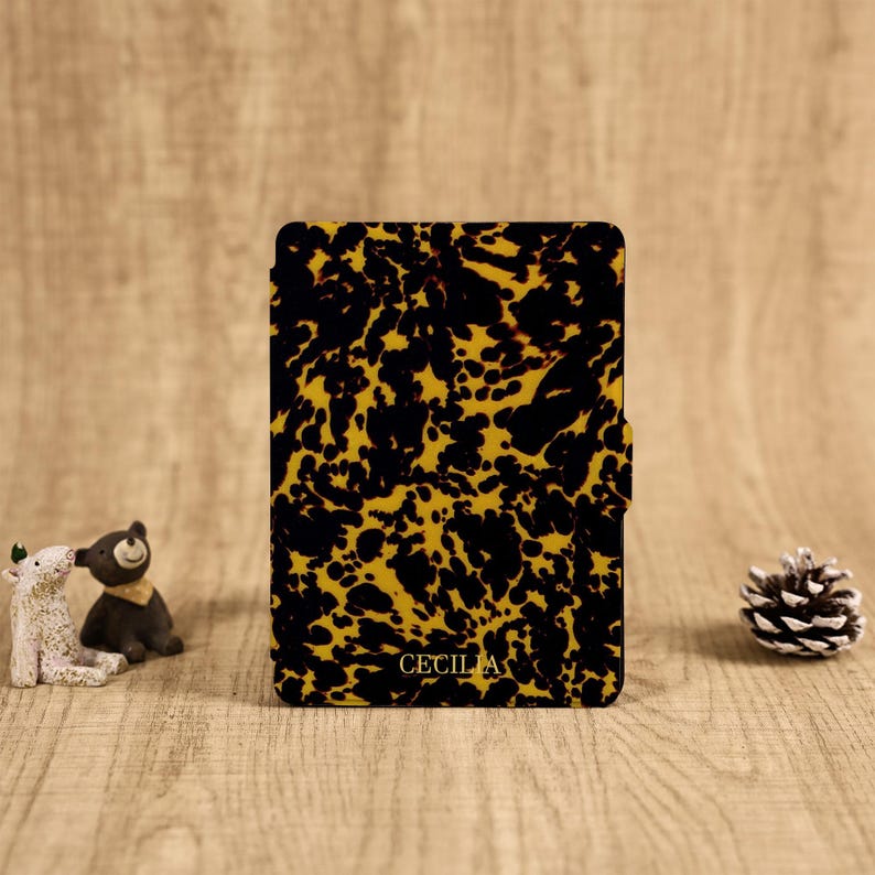 May include: A rectangular e-reader case featuring a black and amber tortoiseshell pattern with the name CECILIA printed in gold lettering at the bottom center. The case has a smooth finish and a small tab on the right side for opening.