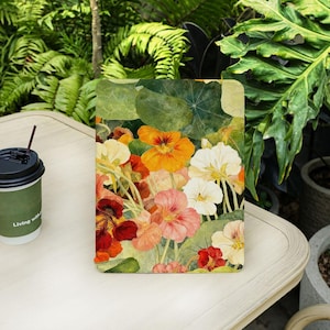 May include: A tablet case with a floral design, showcasing a watercolour painting of nasturtium flowers in orange, red, pink, and white, with green leaves. The case rests on a white table, next to a coffee cup.