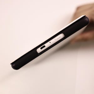 May include: A black protective case for a tablet or electronic device, featuring a white cover. The side profile reveals a precise cutout for a charging port, a small circular microphone hole, and a rectangular power button on the white edge.