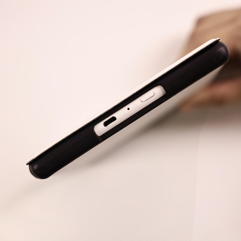 May include: A black protective case for a tablet or electronic device, featuring a white cover. The side profile reveals a precise cutout for a charging port, a small circular microphone hole, and a rectangular power button on the white edge.
