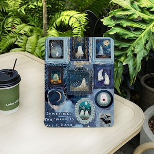 May include: A decorative panel featuring framed illustrations of ghosts, moons, and bats in a blue and white color scheme. The text "Sometimes the moon is all I have" is printed on the panel. A coffee cup with a straw is on the left.