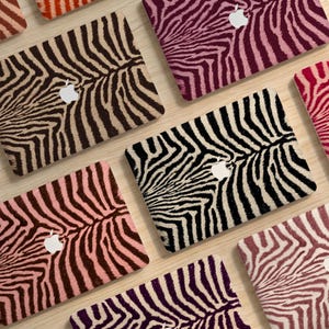 May include: A collection of laptop skins featuring various zebra stripe patterns in colors including black, white, brown, pink, and purple. Each rectangular skin displays a central cutout for an Apple logo, showcasing a textured animal print design.
