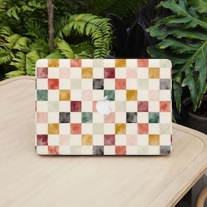 Op de afbeelding: Een laptop met een kleurrijk ruitpatroon in tinten rood, groen, geel en zwart. Het ontwerp is in aquarel stijl, met een wit Apple-logo in het midden. De laptop staat op een licht houten oppervlak.