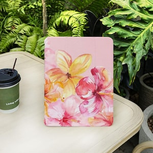 Pode incluir: Uma capa rosa para tablet com um design floral em aquarela. As flores são amarelas, laranja e rosa. A capa está sobre uma mesa clara, com uma xícara de café e vegetação ao fundo.