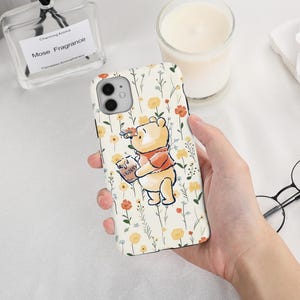 May include: A phone case with a cartoon illustration of Winnie the Pooh holding a honey pot. The case has a floral pattern with yellow and orange flowers on a cream background. The phone case is held in a person's hand.