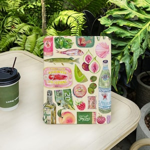 May include: A beige tablet case with a watercolour design of food items such as fish, figs, olives, and a bottle of San Pellegrino. The design also includes a can of Abba sardines and a peach.