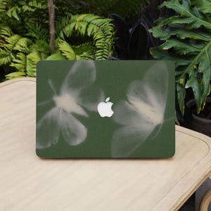 Peut inclure: Une peau d'ordinateur portable verte avec un logo Apple blanc et un motif de deux formes translucides en forme de papillon. La peau d'ordinateur portable est sur une surface en bois de couleur claire, avec un feuillage vert en arrière-plan.