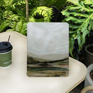 May include: A tablet case featuring an abstract landscape painting in muted tones of green, brown, and gray. The artwork depicts rolling hills and a body of water under a cloudy sky. A coffee cup with the text "Living with" is on the left.