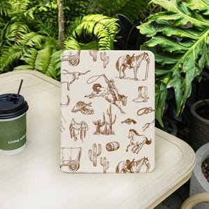 May include: A beige tablet case with a brown cowboy-themed design. The pattern includes illustrations of horses, cacti, a cowboy riding a bucking bronco, and other Western elements. A coffee cup with the text "Living with" is in the foreground.