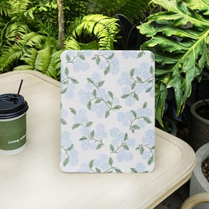Puede incluir: Una funda blanca para tableta con un estampado floral de flores de hortensia azul claro y hojas verdes. La funda está sobre una mesa de color claro junto a una taza de café con una pajita marrón. El fondo incluye follaje verde.