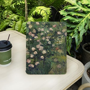 Op de afbeelding: Een rechthoekig notitieboek met een schilderij van een rozentuin in groen, roze en witte tinten. Links staat een koffiekopje met de tekst "Living with".