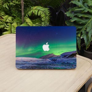 May include: A laptop skin featuring a vibrant night sky with green aurora borealis lights over a snow-covered mountain landscape. The design includes a central cutout for the white Apple logo, set against a deep blue and purple starry sky background.