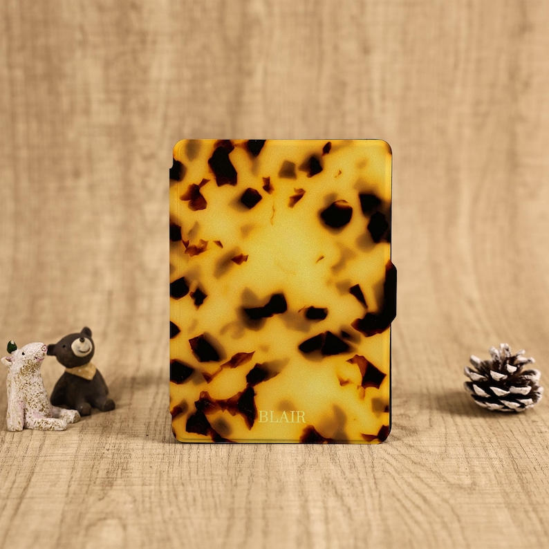 May include: A rectangular electronic reader case featuring a glossy tortoiseshell pattern in shades of amber, brown, and black. The word BLAIR is printed in small, centered capital letters near the bottom edge of the front cover surface.