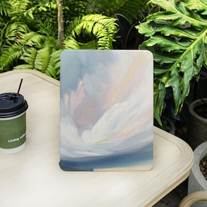 Puede incluir: Una obra de arte rectangular con esquinas redondeadas, que presenta una pintura abstracta en tonos de azul, blanco y melocotón. La obra de arte está colocada sobre una mesa de color claro, con una taza de café visible a la izquierda.