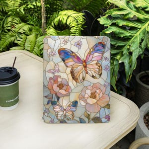 May include: A decorative tablet cover featuring a stained-glass design with a large butterfly and two smaller butterflies, along with several flowers in shades of pink, blue, and yellow. The cover is on a white table with a coffee cup.