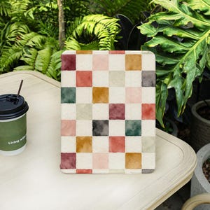 May include: A tablet case with a watercolor checkered pattern in shades of red, pink, green, and gold. The case is on a white table next to a coffee cup with the words "Living with" printed on it.