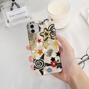 May include: A decorative phone case featuring a cream-colored background with various star shapes in gold, black, and red. The case also includes a small disco ball and a floral design. The phone case is held in a person's hand.