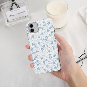 May include: A white phone case with a pattern of small blue flowers is held in a hand. The case has a glossy finish and a camera cutout. A glass bottle of perfume and a lit candle are in the background.