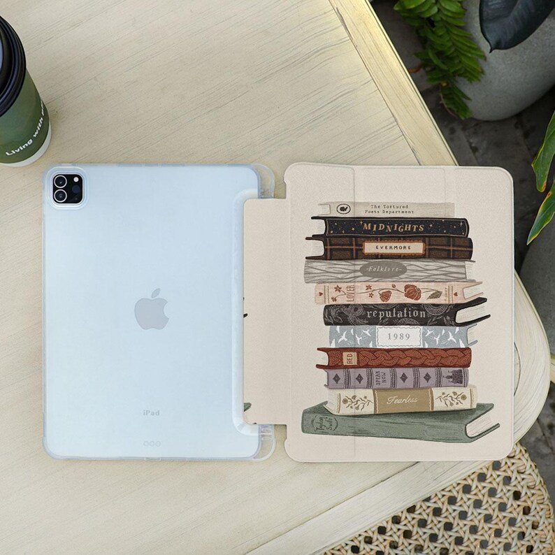 May include: A light blue iPad with a clear case and a cover featuring a stack of books. The book titles include "Midnights", "Evermore", "Reputation", and "1989". The cover has a beige background.