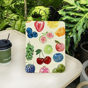 May include: A cream-colored tablet case featuring a watercolor design of various fruits, including strawberries, blueberries, cherries, and oranges. The case is on a light-colored table with a green and brown coffee cup and lush green foliage in the background.