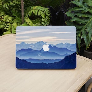 May include: A laptop with a blue mountain range design. The image features a gradient of blue hues, depicting layers of mountains under a cloudy sky. The Apple logo is visible on the laptop.