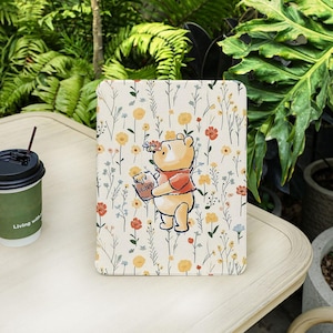 Pode incluir: Uma capa para tablet com um design do Ursinho Pooh. A capa tem um fundo creme com um padrão floral e uma ilustração de desenho animado do Ursinho Pooh segurando um pote de mel. A capa está em uma mesa branca com uma xícara de café.