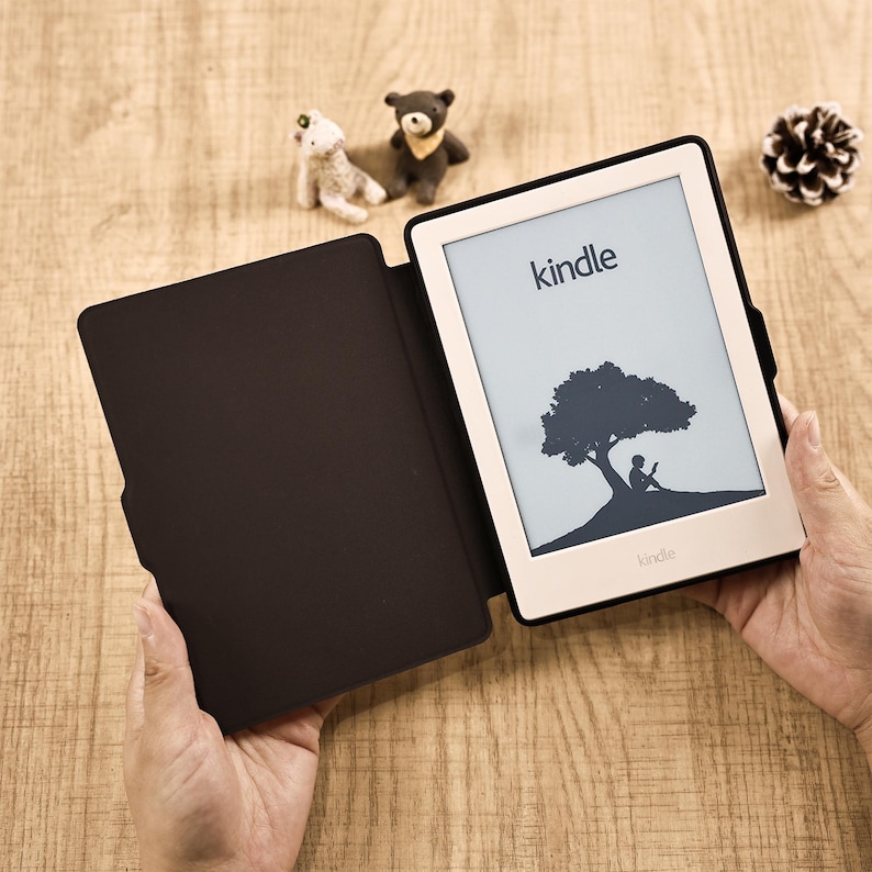 May include: A Kindle e-reader with a light-colored frame and a black protective cover. The screen displays the word kindle at the top and a graphic of a person sitting under a tree reading a book. The device is held open, revealing the dark interior.