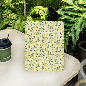 Puede incluir: Una funda rectangular para tableta con un estampado de limones y bayas. El diseño presenta limones amarillos, pequeñas bayas azules y hojas verdes sobre un fondo amarillo claro. La funda está sobre una mesa blanca con una taza de café.