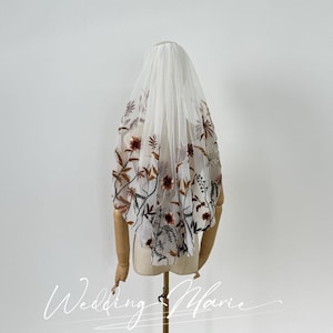 Op de afbeelding: Een witte tule bruids sluier met bloemenborduurwerk in tinten bruin, rood en groen. De sluier is op een mannequin hoofd.