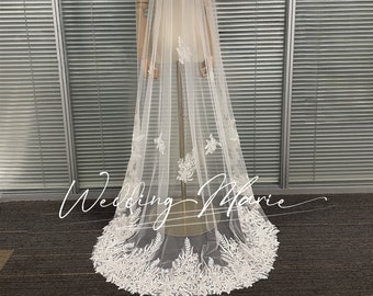 May include: A long, white bridal veil with intricate floral and leaf lace detailing along the bottom edge. The veil is displayed on a mannequin, showcasing its length and delicate design. The text "Wedding Marie" is written on the veil.