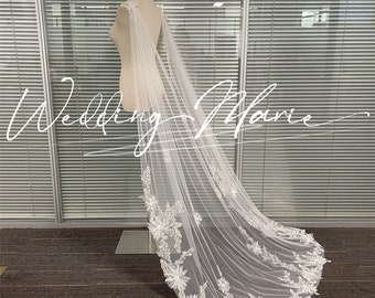 May include: A white tulle wedding veil with delicate floral lace and bead embellishments. The veil is draped over a mannequin and has a long train.